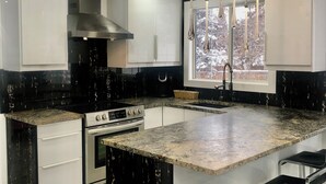 Una cocina moderna con armarios blancos y negros, una isla central con taburetes de bar, electrodomésticos de acero inoxidable y un ventanal con vistas a árboles.