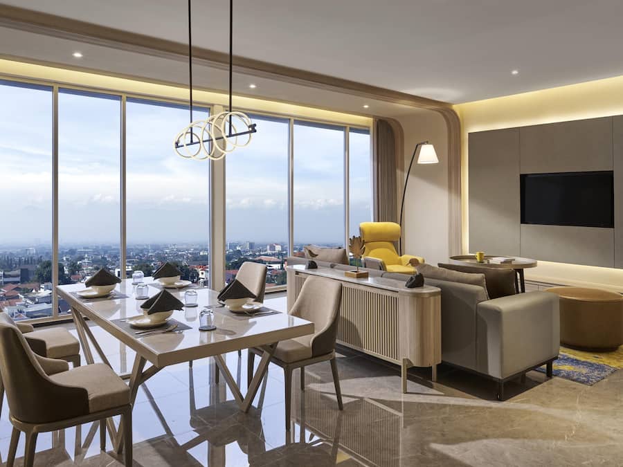 A modern hotel room with a large dining table, comfortable seating, a sofa, a TV mounted on the wall, and a city view through large windows.