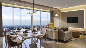 Una habitación de hotel moderna con una amplia mesa de comedor, asientos cómodos, un sofá, un televisor montado en la pared y vistas a la ciudad a través de grandes ventanales.