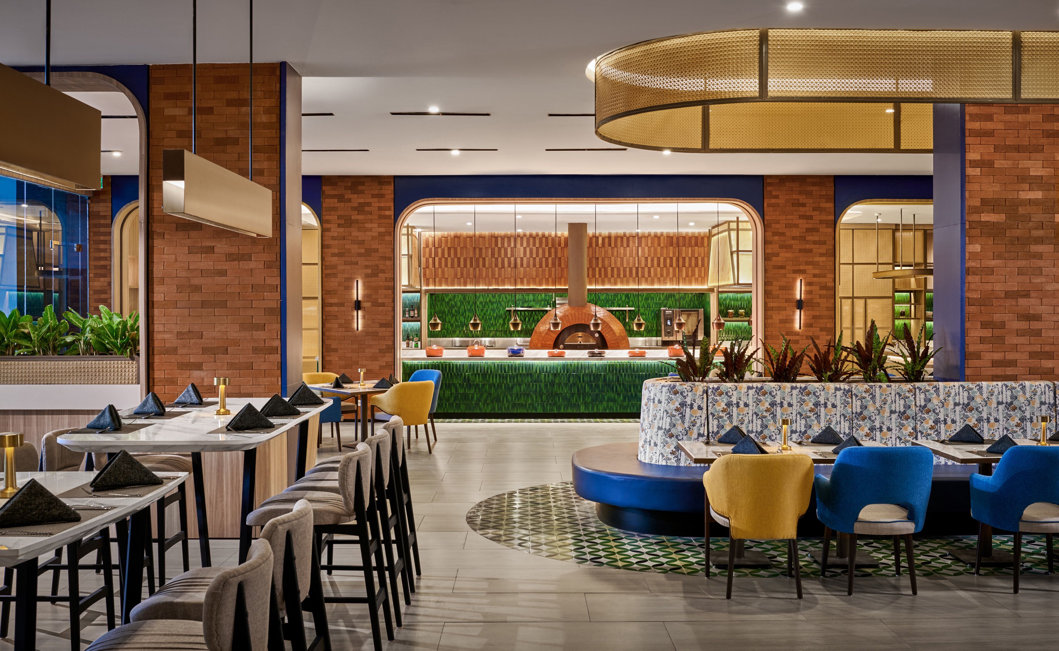 A modern restaurant with brick walls, a green wall with a kitchen, and a seating area with blue and yellow chairs.