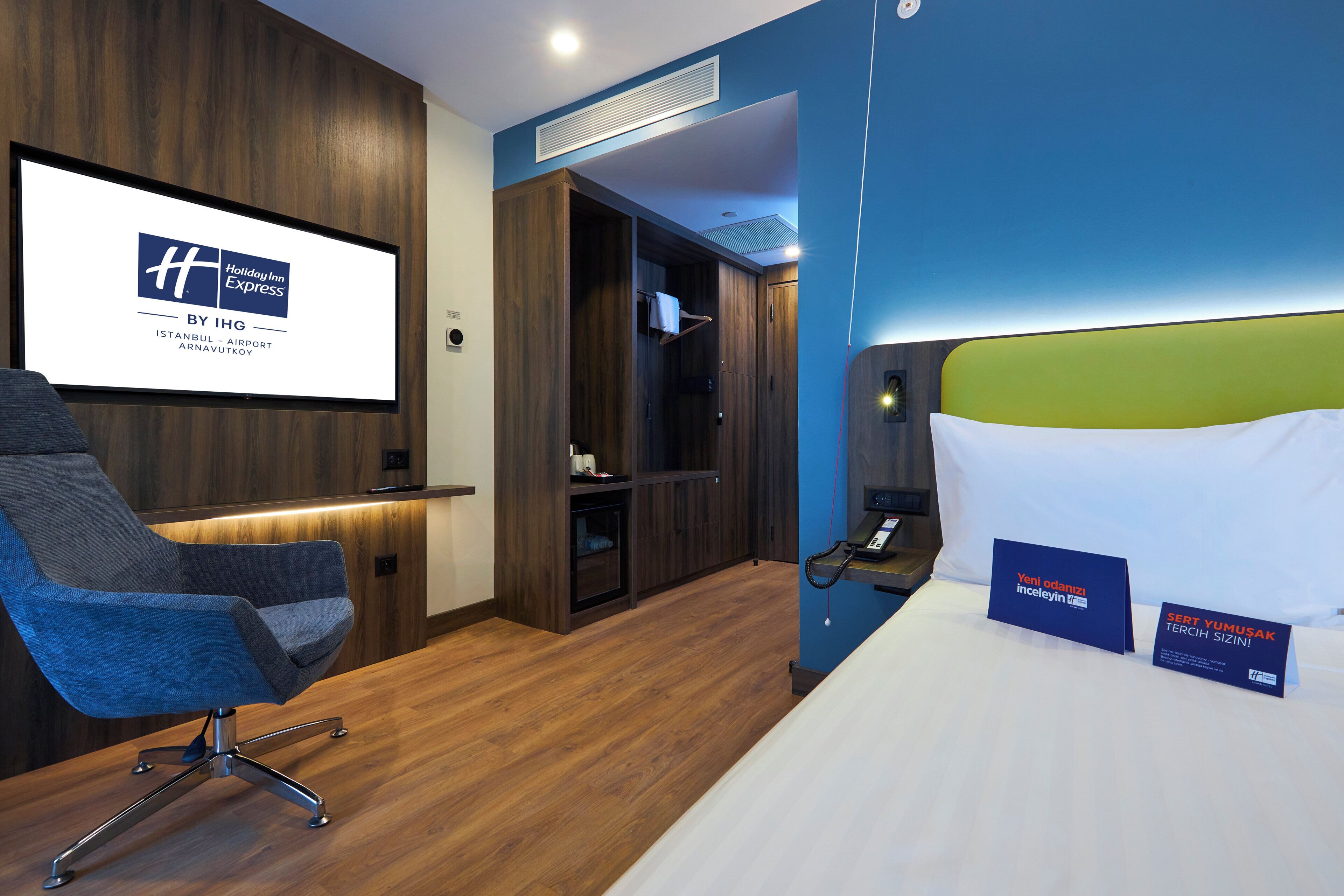 Una camera d'albergo con pavimento in legno, una scrivania con sedia, un letto con testiera verde e uno schermo a parete che mostra il nome dell'hotel.