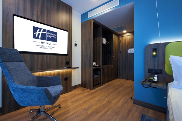 Habitación de hotel con piso de madera, pared azul, cama, escritorio, silla y pantalla con el logotipo de Holiday Inn Express.
