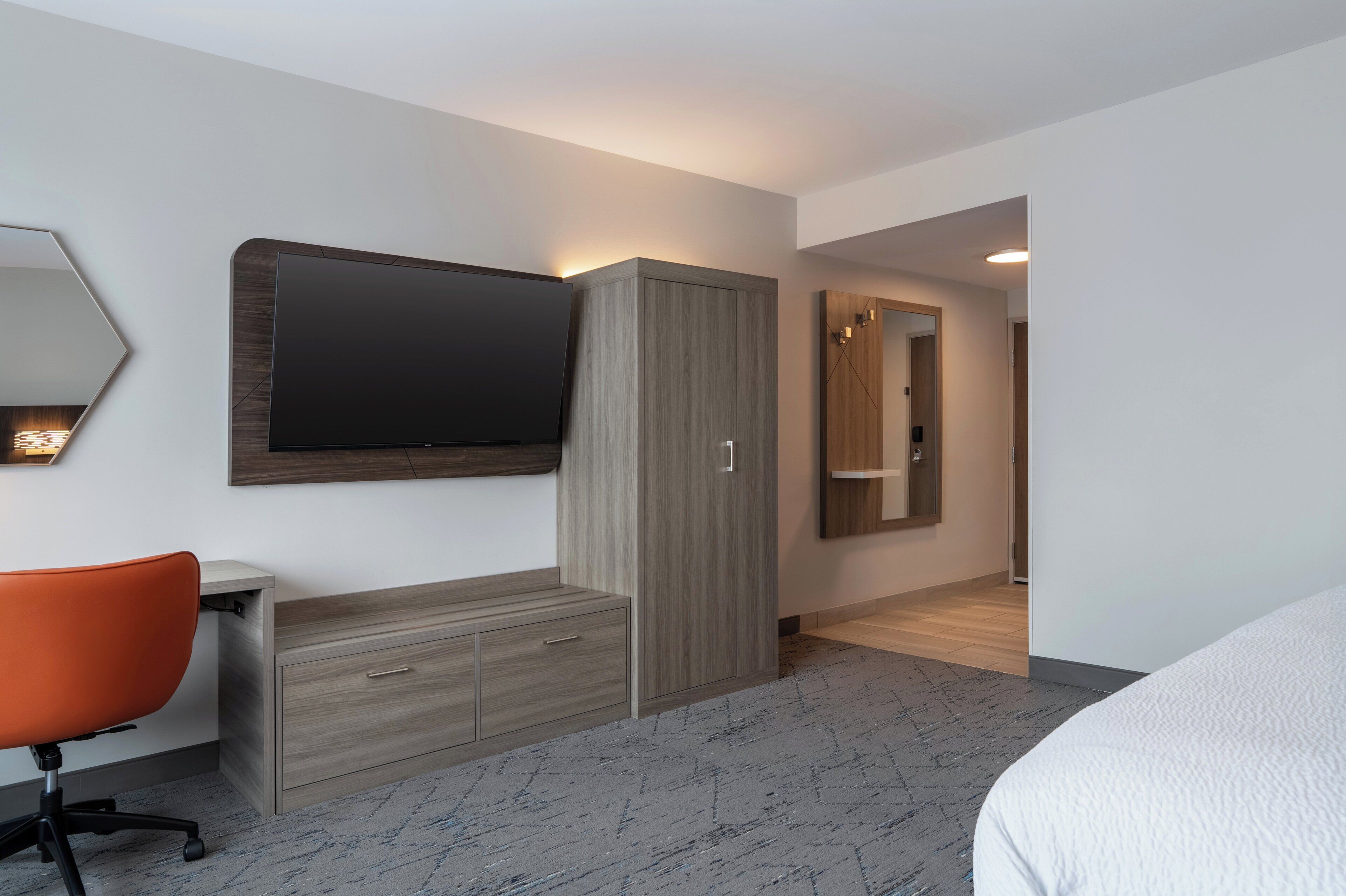 Ein modernes Hotelzimmer mit einem Flachbildfernseher, einem Holzschrank, einem Schreibtisch mit orangefarbenem Sessel und einem großen Bett.