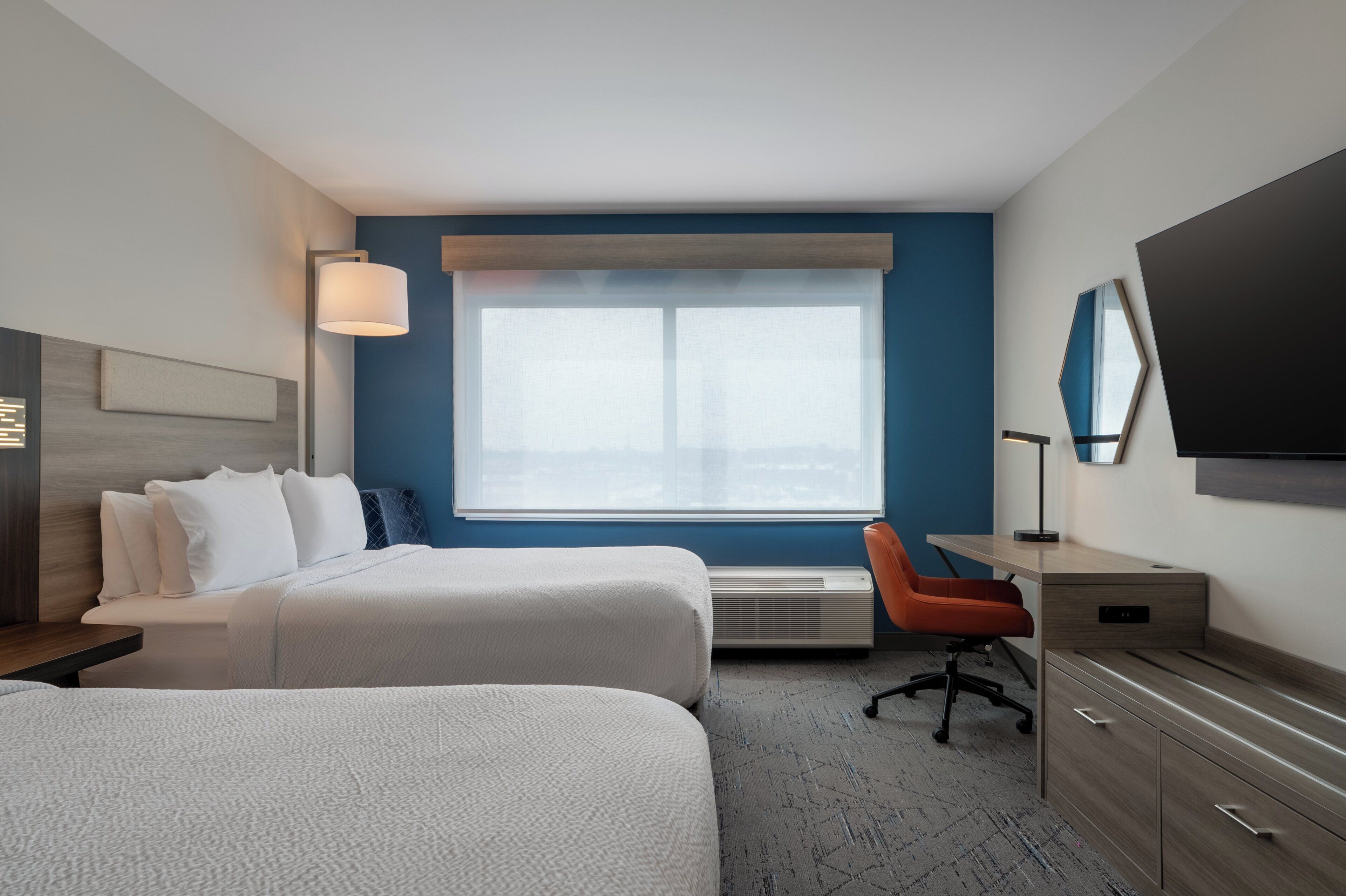A hotel room with two beds, a desk, an orange chair, a flat-screen TV, and a large window.