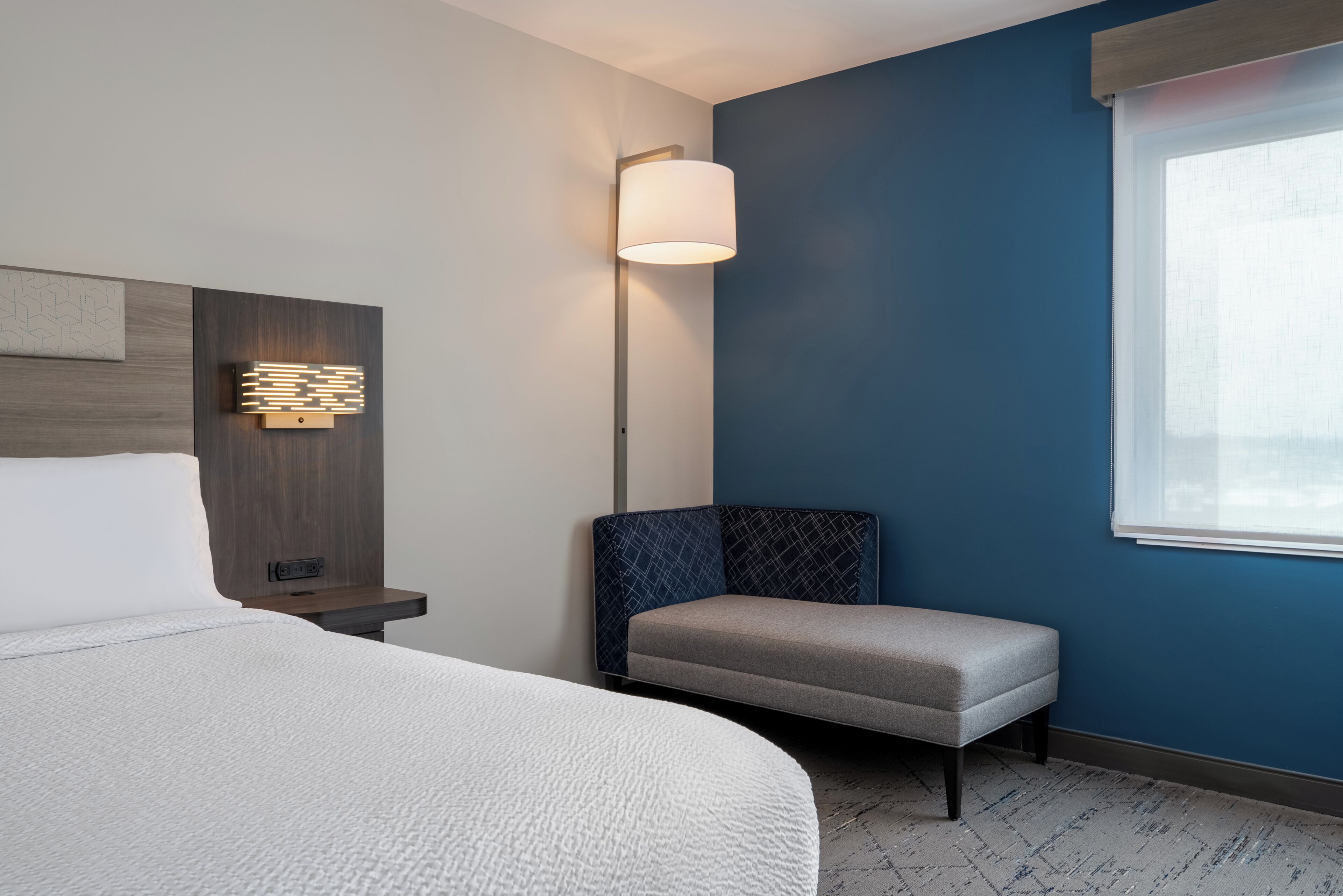 Ein Hotelzimmer mit Bett, Nachttisch, einer Wandleuchte, einer Bank und einem Fenster mit Jalousien.