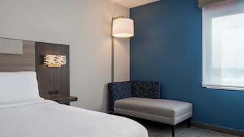 Ein Hotelzimmer mit Bett, Nachttisch, einer Wandleuchte, einer Bank und einem Fenster mit Jalousien.