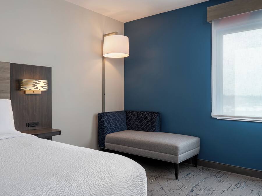Ein Hotelzimmer mit Bett, Nachttisch, einer Wandleuchte, einer Bank und einem Fenster mit Jalousien.