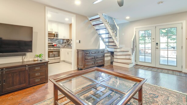 A spacious living room with a large flat-screen TV, a glass coffee table, and a dark wooden entertainment center. A staircase with a glass railing leads to an upper level. - Near Hiking & Golf: Cozy Cottage in Morgan Hill! (Morgan Hill)