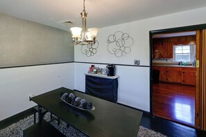 Un espace repas comprenant une table, des chaises, un lustre, un élément décoratif mural et une vue sur la cuisine.