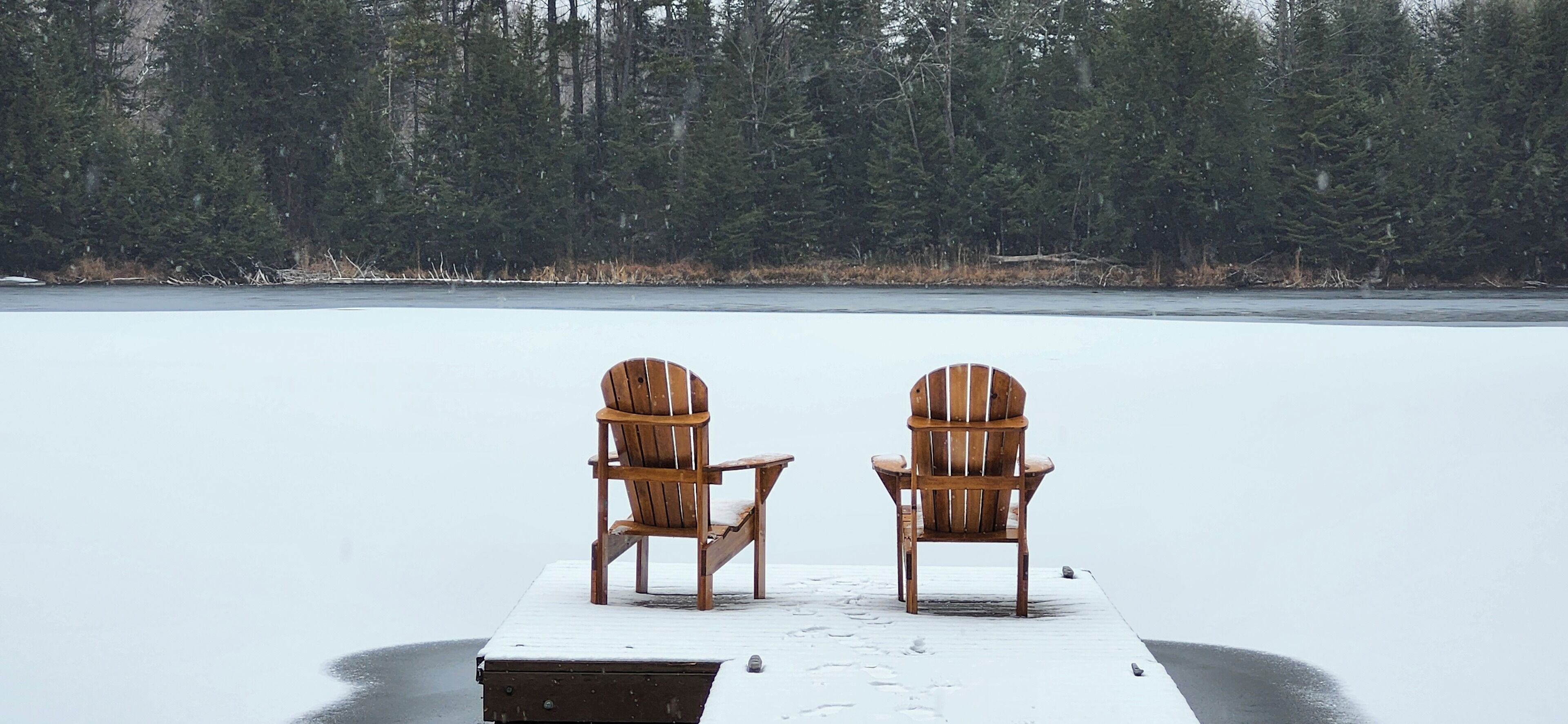 Two wooden Adirondack chairs on a dock extending into a frozen lake, surrounded by dense forest.