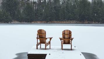 Two wooden Adirondack chairs on a dock extending into a frozen lake, surrounded by dense forest.