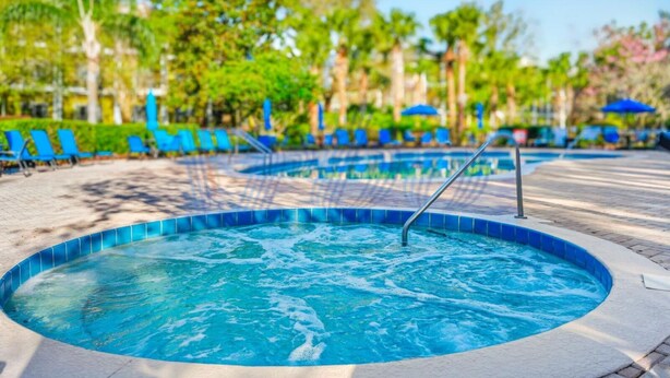 Un jacuzzi con borde de azulejos, rodeado de sillones de descanso y sombrillas, con árboles y un cielo despejado al fondo.