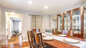 Un comedor con una mesa de madera dispuesta para cuatro personas, sillas, un armario con puertas de vidrio y una cortina estampada.