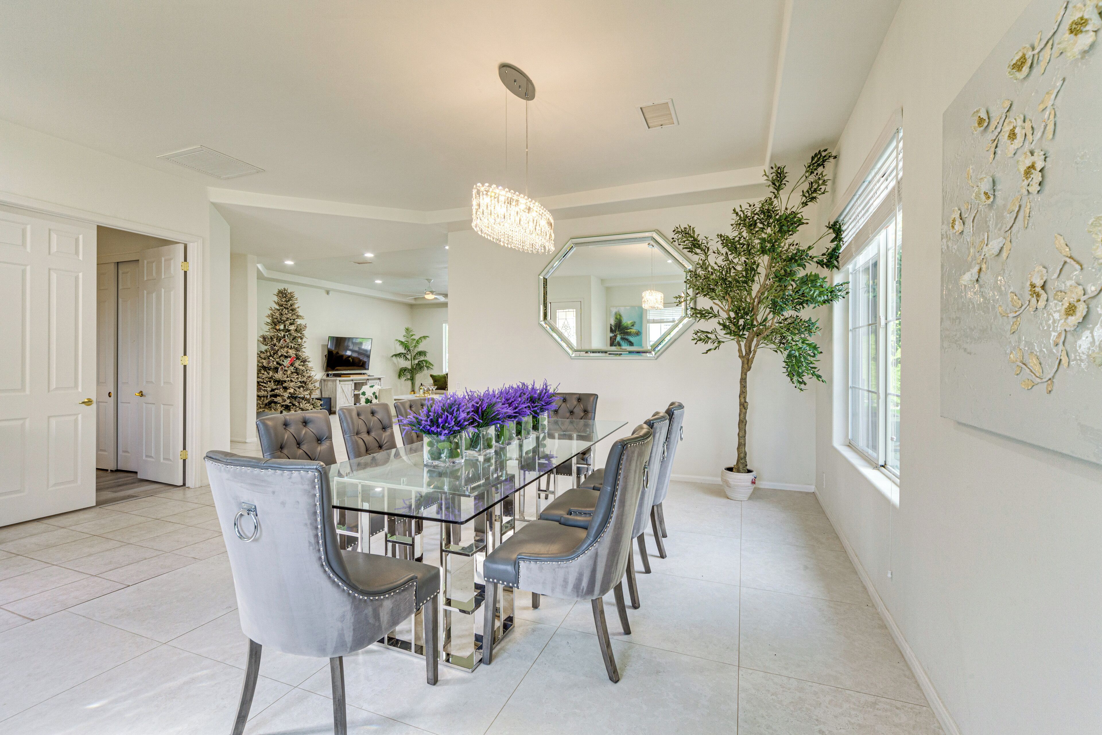 A dining area with a glass table, grey upholstered chairs, a chandelier, and a large mirror on the wall.