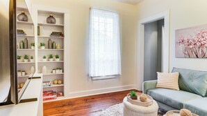 A living room with a bookshelf, a sofa, a window with sheer curtains, and a painting on the wall. - Walkable Location: Quaint Cottage in Forest Grove! (Forest Grove)