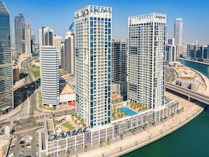 A high-rise building with a prominent 'DAMAC' sign, surrounded by other modern high-rises, a waterfront, and a busy road.