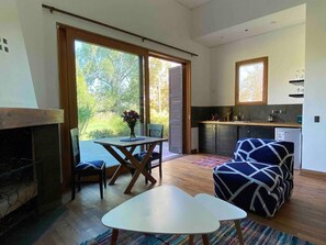 Una sala de estar moderna con chimenea, una mesa de comedor y un sofá. Se observa una zona de cocina con ventana y una pequeña isla.