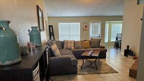 A living room with a sectional sofa, a coffee table, and a large vase. There are two windows with blinds, a door, and a small table with a computer.
