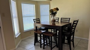 A dining room with a wooden table and four chairs, a vase with flowers on the table, and two windows with blinds.