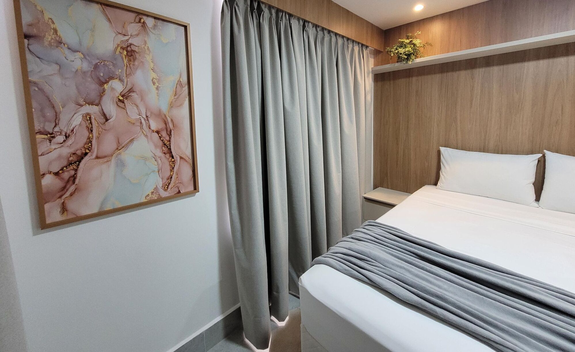 A modern hotel room with a large bed, grey curtains, a wooden headboard, and a marble artwork on the wall.