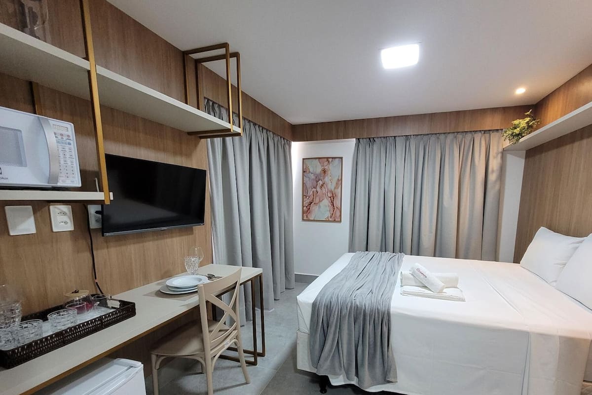 Quarto de hotel com cama, escrivaninha com televisão de tela plana, mesinha com bandeja de copos e um quadro na parede.