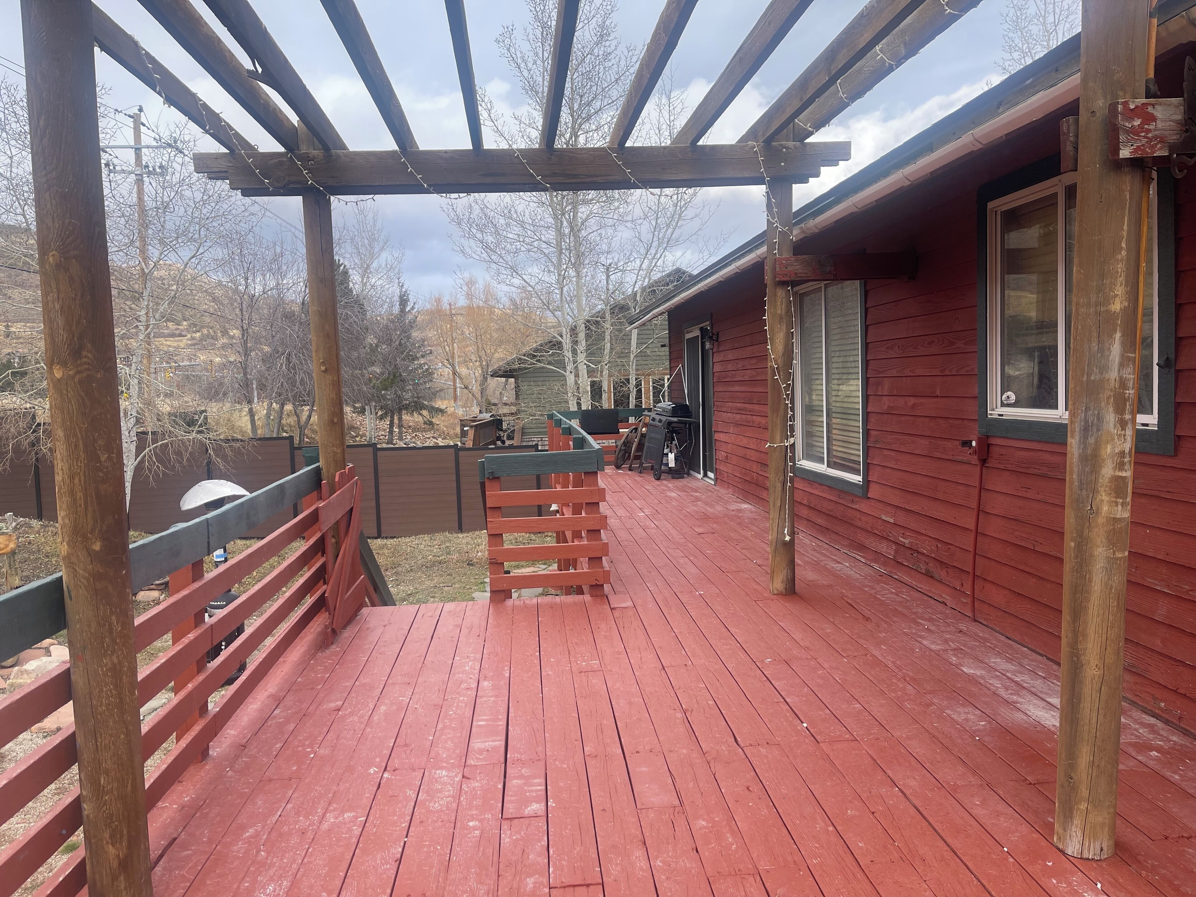 Une terrasse en bois avec une pergola, un bâtiment rouge doté de fenêtres et un espace clos aménagé avec du mobilier d’extérieur.
