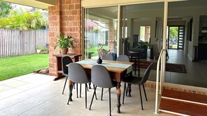 Eine Terrasse im Freien mit einem Holztisch und Stühlen, einer Ziegelwand und einer Glasschiebetür, die in einen überdachten Bereich führt.