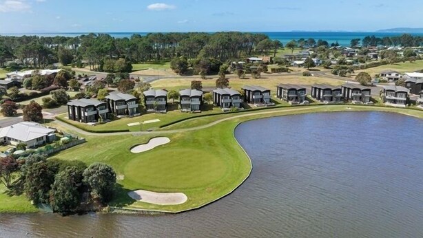 A golf course with a water hazard, surrounded by residential buildings and a clear sky. - Dunes Lake View Apartment (Matarangi)