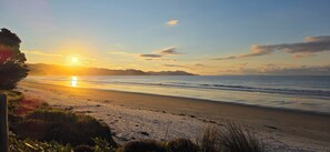 A beach with a sunset, waves, and distant mountains. - Dunes Lake View Apartment (Matarangi)