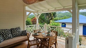 Un área de estar techada al aire libre con una mesa preparada para comer, rodeada de barandas blancas y vistas a un paisaje exuberante.