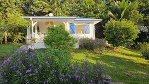 Un pequeño edificio blanco con contraventanas azules, rodeado de vegetación exuberante y flores moradas.