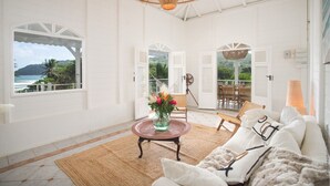 Une pièce spacieuse dotée d’une grande fenêtre offrant une vue sur la mer et les montagnes, d’un luminaire suspendu en osier et d’un coin salon confortable avec un canapé et une table agrémentée d’un vase de fleurs.