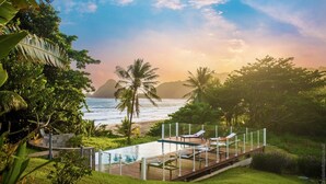 Un espace aménagé au bord de la piscine, avec des chaises longues, une terrasse en bois et une vue sur l’océan et les montagnes.