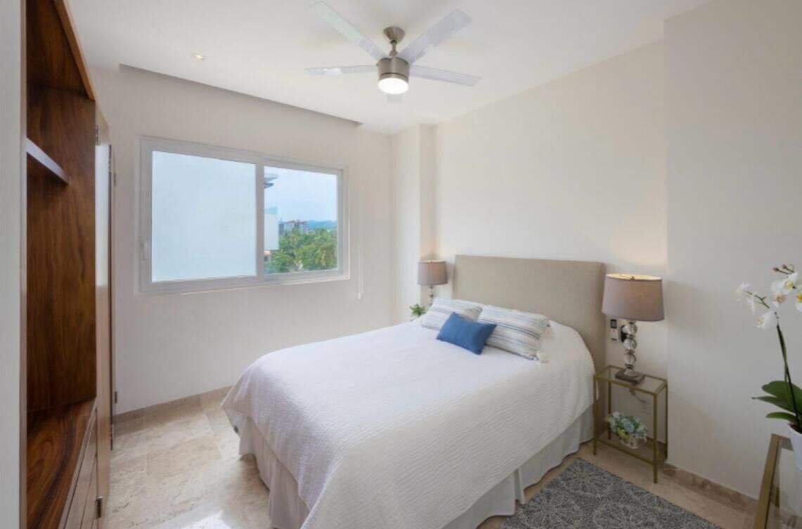 Ein Schlafzimmer mit Bett, Deckenventilator, Fenster und Nachttisch mit Lampe.