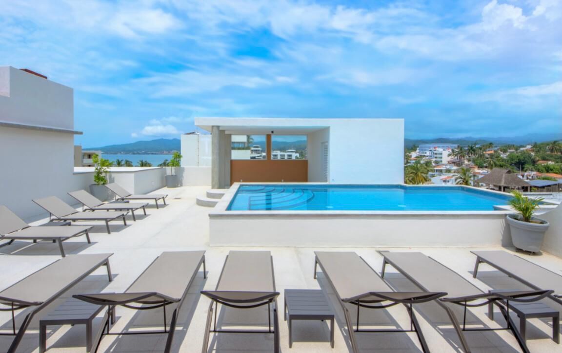 A rooftop pool area with sun loungers and a view of the city and mountains.