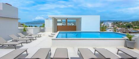 A rooftop pool area with sun loungers and a view of the city and mountains.