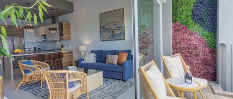 Een moderne woonkamer met een blauwe bank, rieten stoelen, een salontafel en een glazen wand met een kleurrijke muurschildering.
