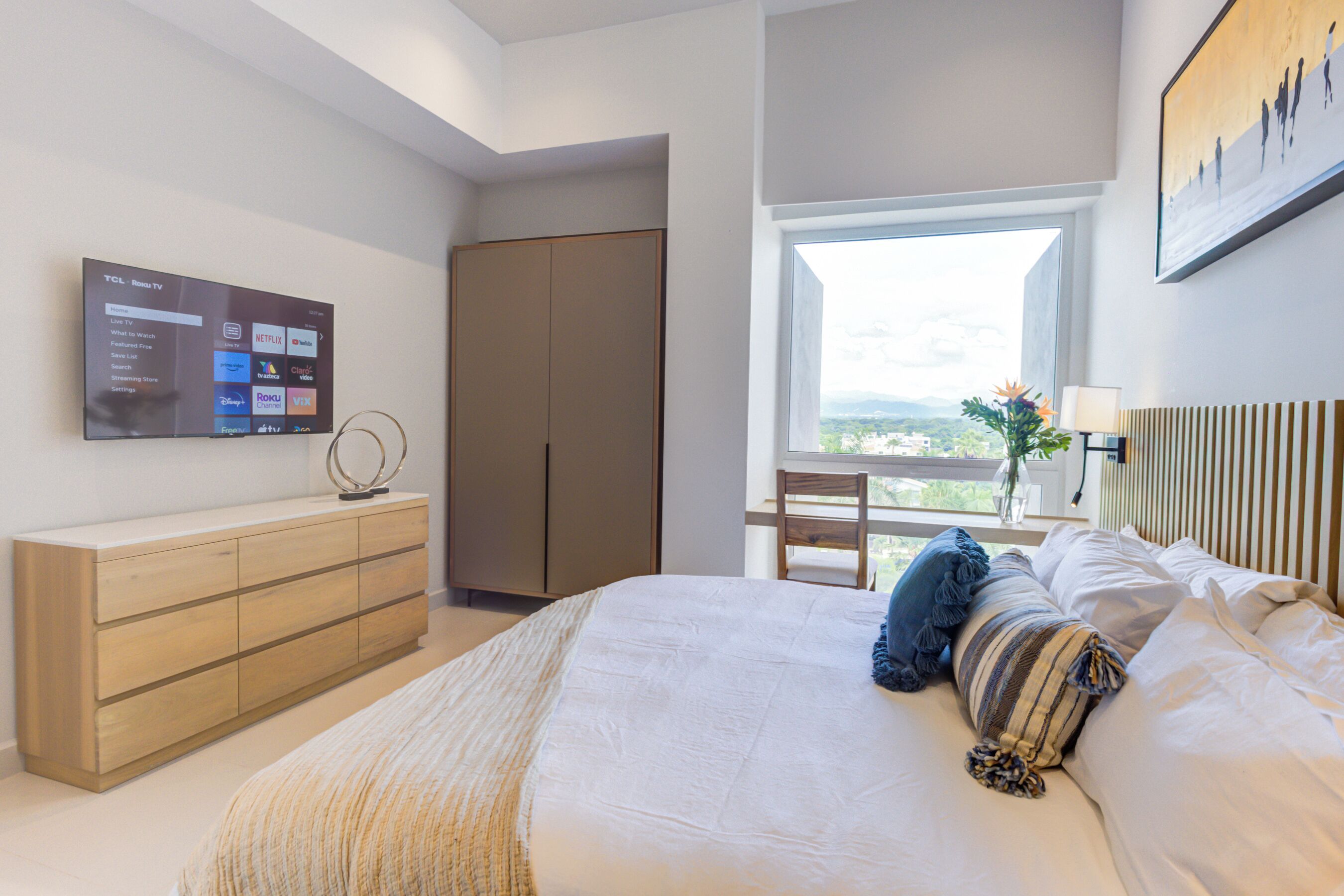 A modern bedroom with a bed, a TV mounted on the wall, a wardrobe, and a window with a view.