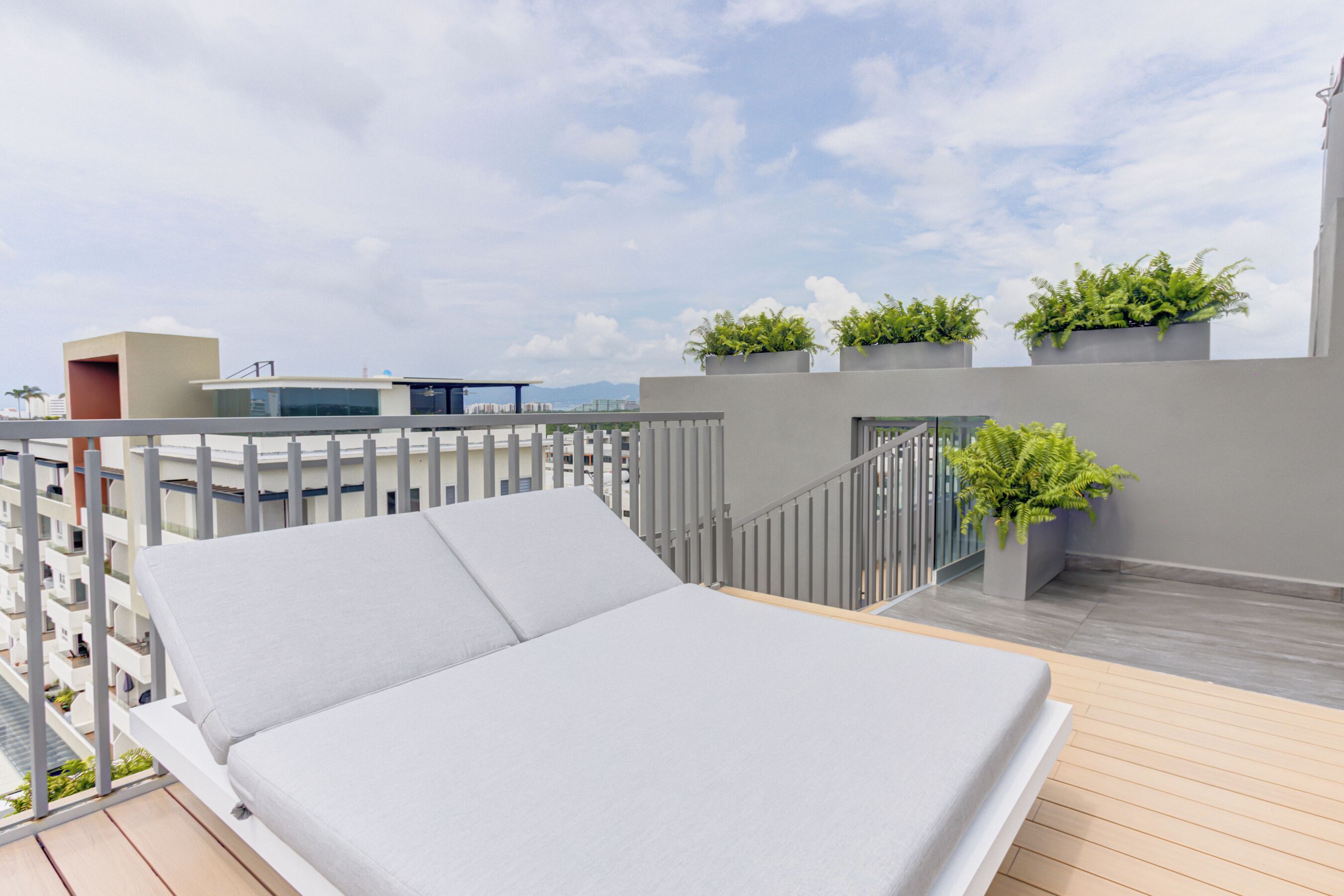 A rooftop lounge with a white cushioned sofa, grey railings, and potted plants.