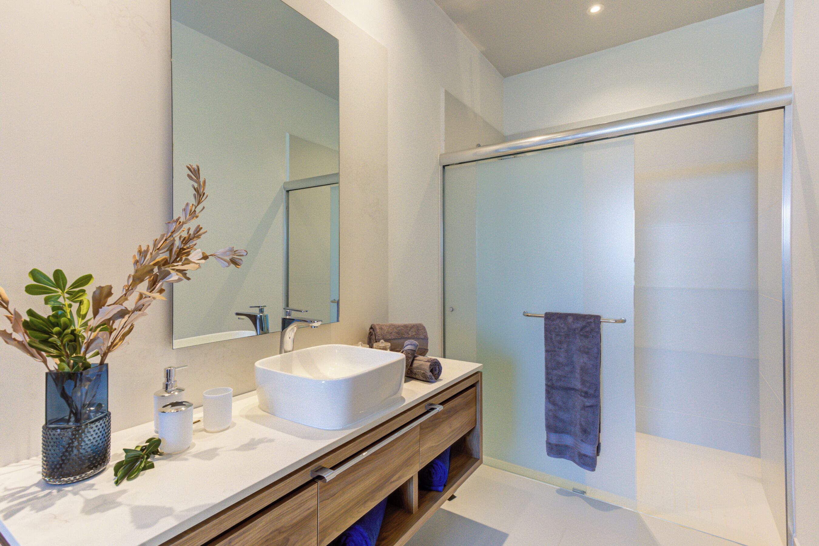 Un baño moderno con un espejo grande, un lavamanos blanco, cabina de ducha de vidrio y tocador de madera.