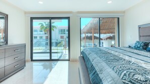 Un dormitorio con una cama grande, un aparador y un ventanal corredizo que da a un balcón con vistas a edificios y una palmera.