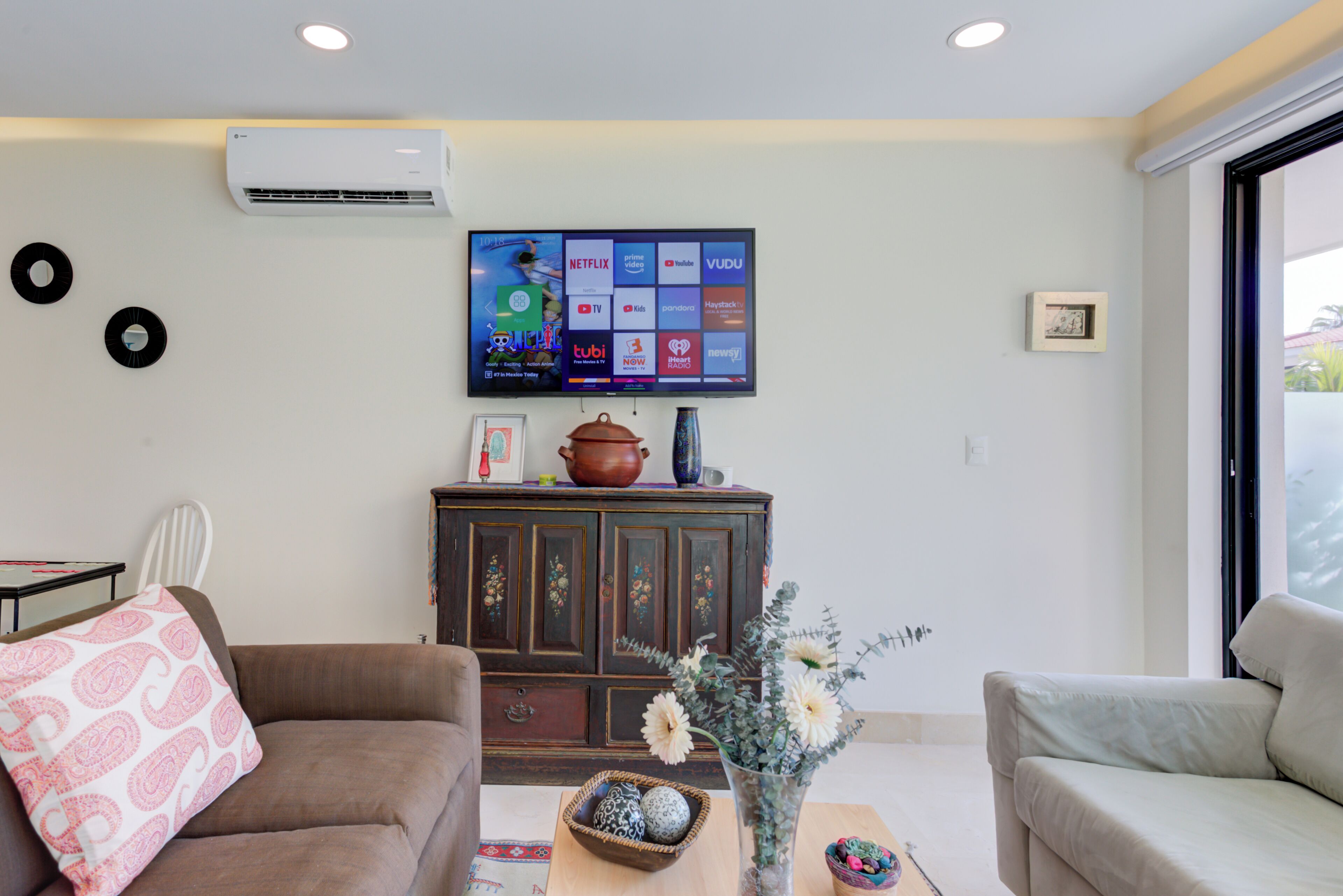 Een woonkamer met een flatscreen televisie aan de wand, een bruine bank, een beige bank, een salontafel met decoratieve items en een kast met een vaas en een pot.