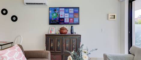Een woonkamer met een flatscreen televisie aan de wand, een bruine bank, een beige bank, een salontafel met decoratieve items en een kast met een vaas en een pot.