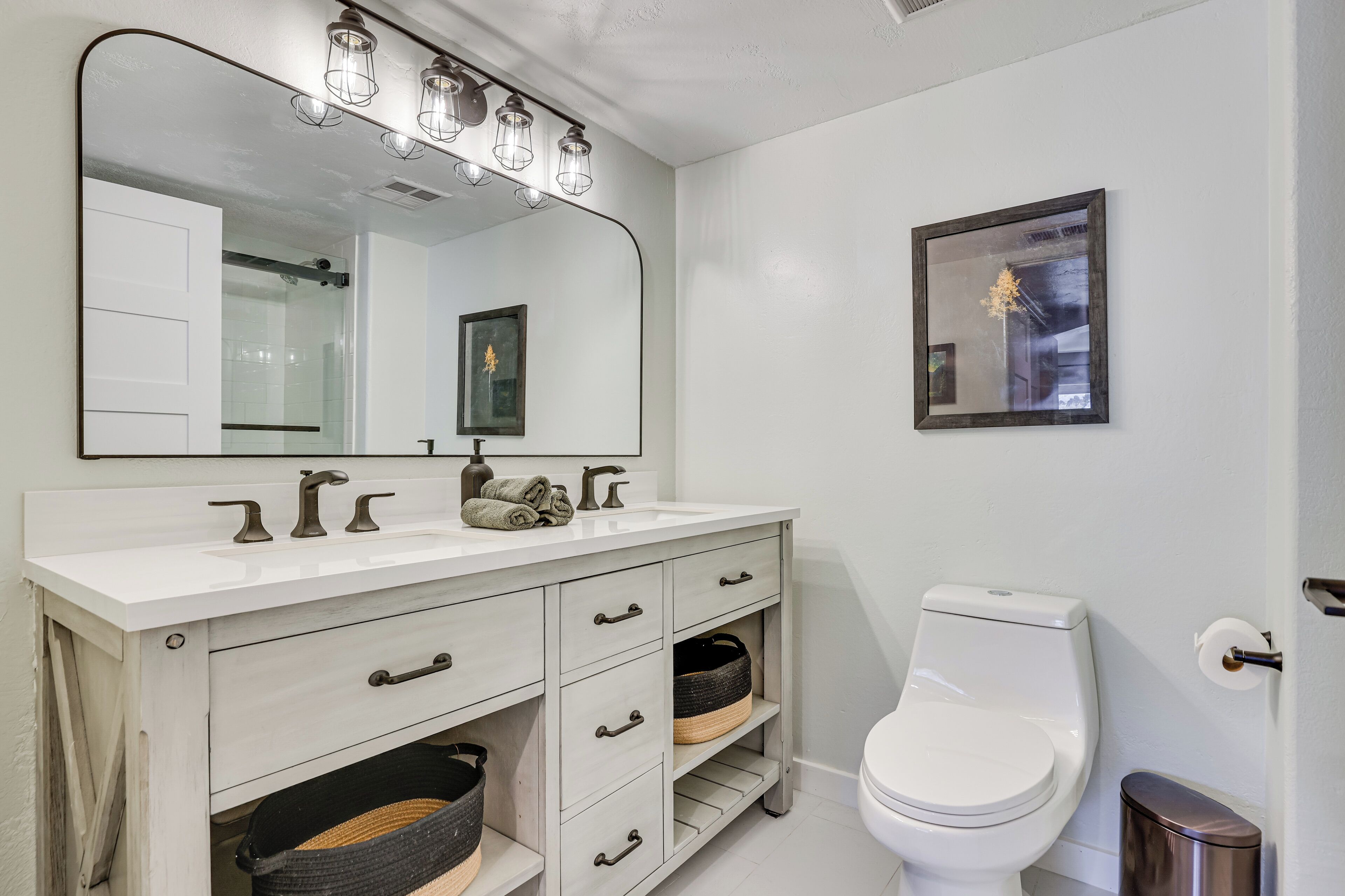 A bathroom with a white vanity, a large mirror with lights, a toilet, and a framed picture on the wall.