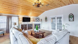 Een ruime woonkamer met een houten plafond, een open haard, een televisie en een groot raam met gordijnen.