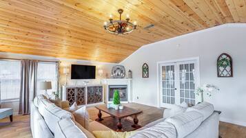 Un salon spacieux doté d’un plafond en bois, d’une cheminée, d’un téléviseur et d’une grande fenêtre avec des rideaux.