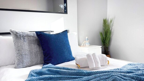 A neatly made bed with a blue blanket, two pillows, and a small bedside table with a bottle and a plant. - Rhythm & Blues Retreat near Syracuse University! (Syracuse)