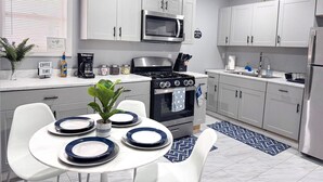 A modern kitchen with white cabinets, a stainless steel stove, a microwave, and a refrigerator. There is a dining table set with plates and a potted plant. - Rhythm & Blues Retreat near Syracuse University! (Syracuse)