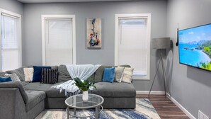 A modern living room with a grey sectional sofa, a glass coffee table, and a large flat-screen TV on the wall. - Rhythm & Blues Retreat near Syracuse University! (Syracuse)