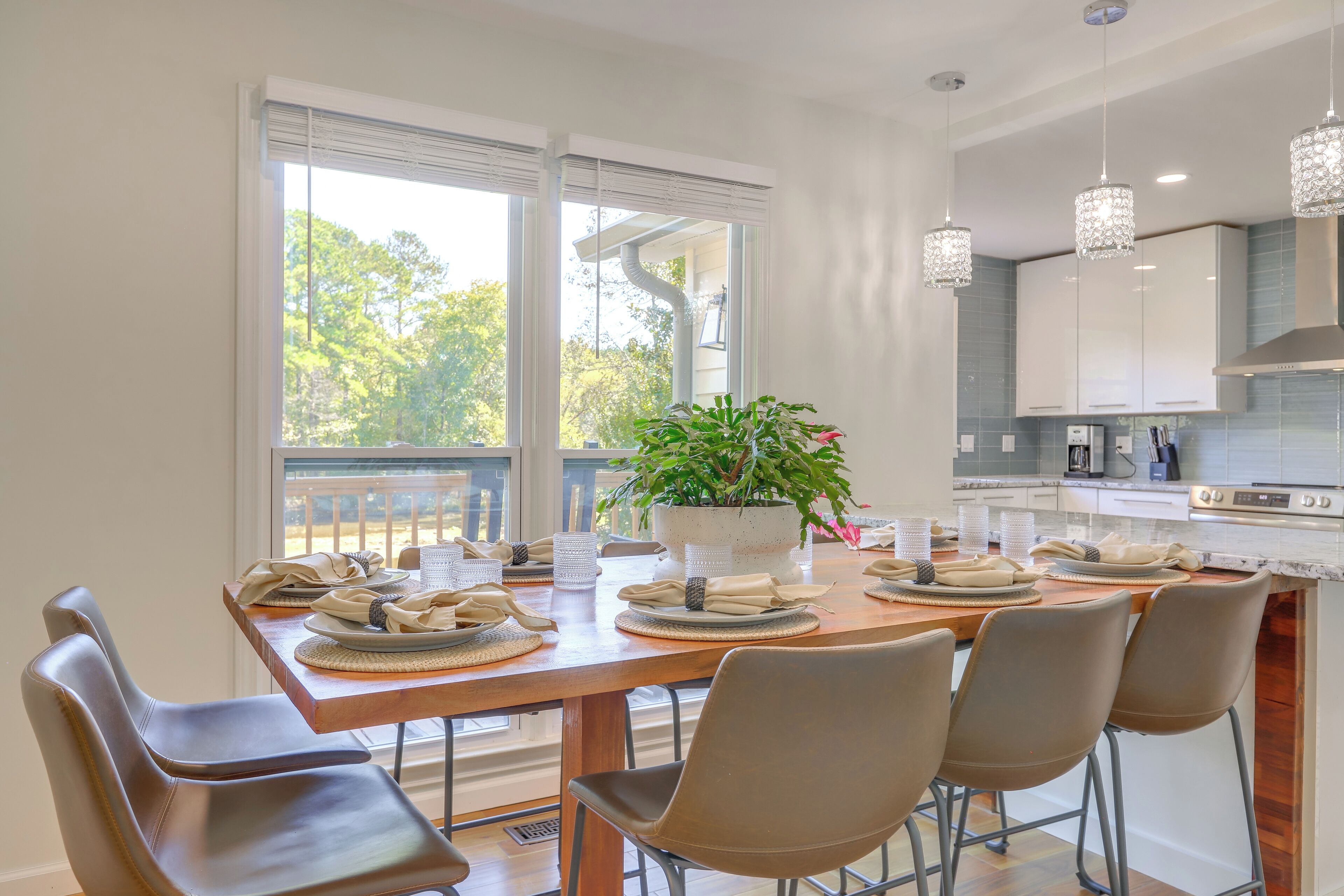 Un coin repas avec une table en bois dressée pour six personnes, une plante en pot et une vue sur l’extérieur grâce à de grandes fenêtres.